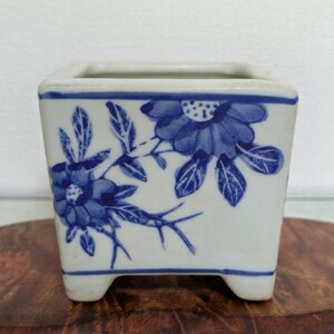 May include: A square, white ceramic planter with a blue floral design. The planter has a blue rim and is supported by four small feet. The floral pattern includes detailed blue flowers and leaves, creating a classic, decorative look.