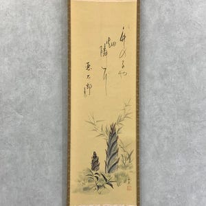 May include: A vintage Japanese scroll painting featuring bamboo shoots and foliage in shades of green and gray. The artwork includes calligraphy in black ink and is framed with a brown border.