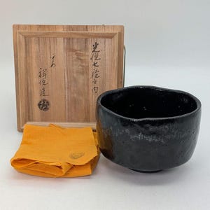 May include: A black ceramic tea bowl with a textured surface, presented with a wooden box and a folded orange cloth. The wooden box has Japanese characters on the front.