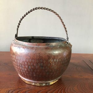 May include: A copper-toned metal bucket with a twisted handle. The bucket features a textured pattern on its lower half and rests on a wooden surface. The handle is secured with metal brackets.