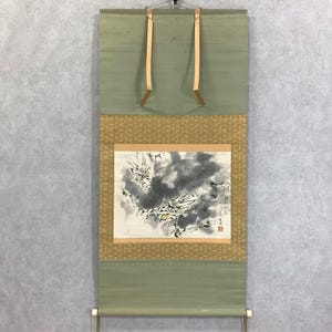 May include: An antique Japanese scroll painting with a nature scene in black and gray ink. The scroll has a green background with a gold patterned border and tan leather hanging straps. The painting is mounted on a wooden dowel.