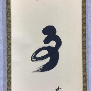 May include: A vertical scroll featuring a large, stylized black calligraphy symbol on a cream-colored background. The symbol has a flowing, brushstroke appearance. The scroll is framed with a decorative border and a light blue fabric.