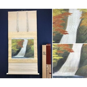 May include: A Japanese scroll painting showing a waterfall scene with autumn foliage. The artwork features a waterfall cascading down a rocky cliff, surrounded by trees with red and yellow leaves. The scroll is cream-coloured with decorative silver accents.