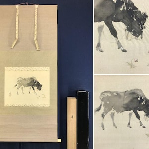 Peut inclure: Un rouleau suspendu présentant une peinture à l'encre de style sumi-e représentant une vache sombre broutant près de petites fleurs. Le rouleau a une bordure en tissu beige clair, des rubans à motifs et un rouleau en bois, avec une boîte.