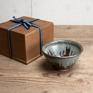 May include: A ceramic bowl with a blue and brown glaze, next to a wooden box tied with a blue and white ribbon. The bowl has a gold rim and a unique, dripped glaze pattern.