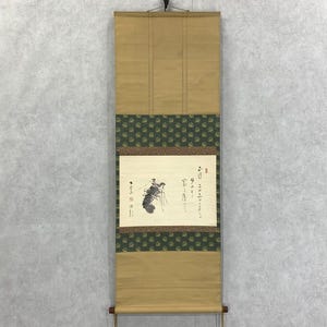 Op de afbeelding: Een traditioneel Japans rolschilderij met een beige achtergrond en een groene patroonrand. Het kunstwerk toont een zwart-wit illustratie van een wezen en kalligrafie. De rol is opgehangen aan een zwarte haak.