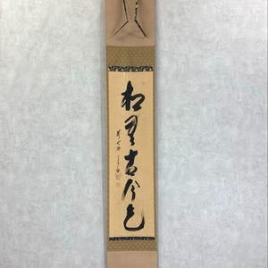 May include: A vertical Japanese scroll with calligraphy in black ink on a beige background. The scroll has decorative gold and brown patterned borders and a wooden top and bottom. The scroll is hanging from a chain.