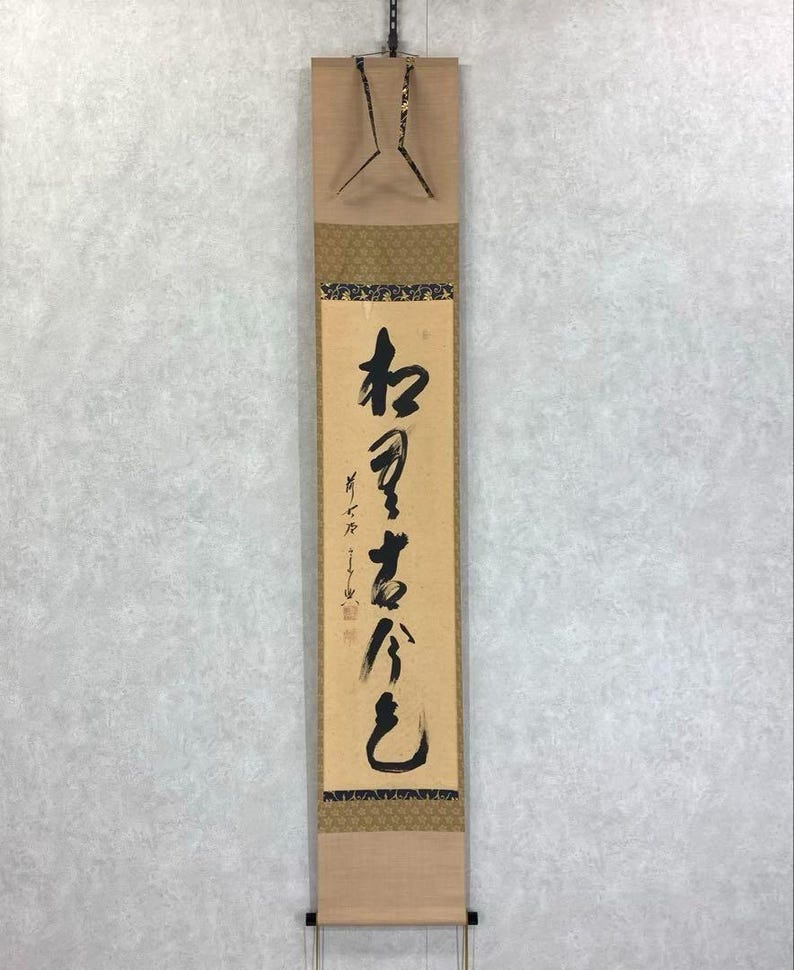 May include: A vertical Japanese scroll with calligraphy in black ink on a beige background. The scroll has decorative gold and brown patterned borders and a wooden top and bottom. The scroll is hanging from a chain.