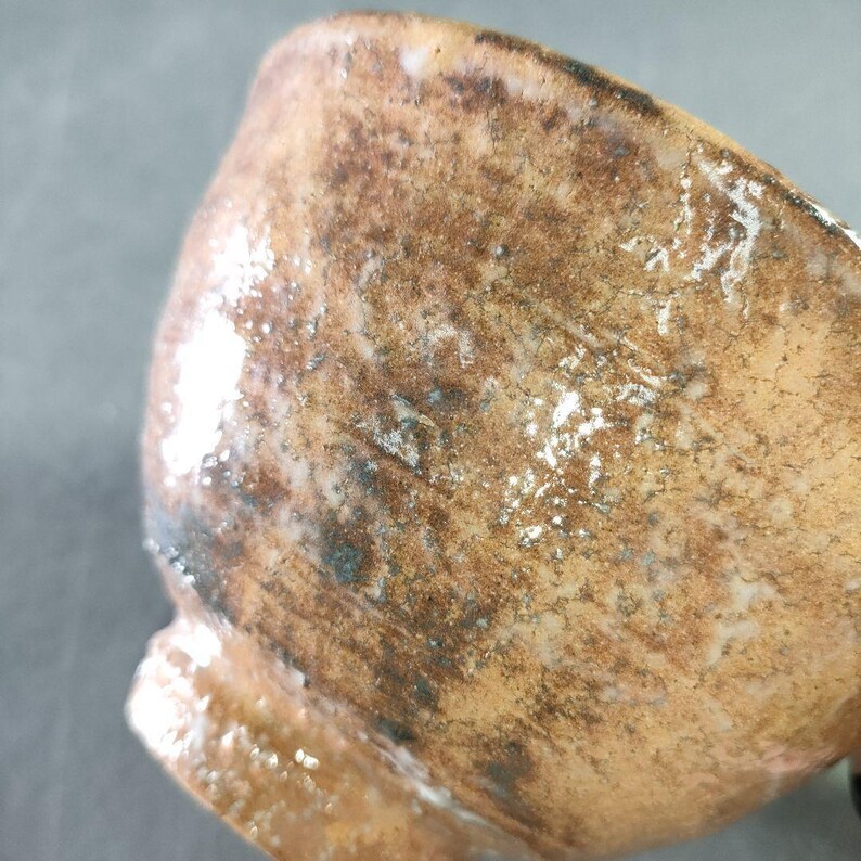 May include: Close-up of a small, handmade ceramic cup. The cup has a textured, earthy brown surface with darker brown and black accents. White speckles add highlights to the glaze. The cup's rim is slightly irregular, suggesting a handcrafted origin.