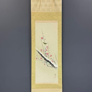 May include: A vertical hanging scroll featuring a painting of a small green bird perched on a snow-covered branch with red plum blossoms. The artwork is mounted on a light gold fabric border with a patterned top section and a wooden roller at the base.