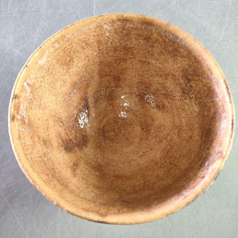 May include: A handmade ceramic bowl with a textured, earthy appearance. The interior features a blend of brown and tan hues, with subtle variations in color and texture. The bowl's rim is slightly lighter in color, creating a visual contrast.