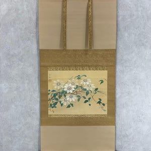May include: A traditional Japanese scroll painting featuring a floral design with white flowers, green leaves, and a light yellow background. The scroll has a beige fabric with gold accents and a black hanging rod.