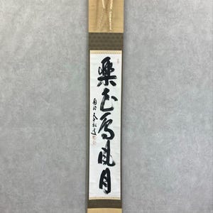Könnte beinhalten: Eine vertikale japanische Schriftrolle mit schwarzer Kalligraphie auf weißem Hintergrund. Die Schriftrolle hat eine beige Ober- und Unterseite mit einem braunen, gemusterten Abschnitt in der Mitte. Der Text ist in vertikalen Spalten geschrieben.