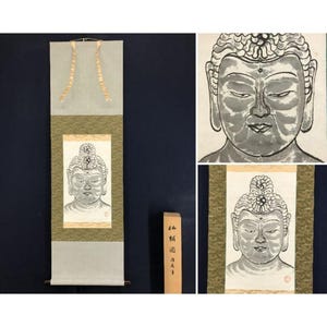 Op de afbeelding: Een traditionele Japanse rolschildering met een gedetailleerde illustratie van het gezicht van Boeddha. Het kunstwerk is voornamelijk in grijstinten en zwart, met een serene uitdrukking. De rol heeft een lichtgroene rand en een houten rol aan de onderkant.