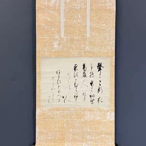 May include: A vertical hanging scroll featuring a central rectangular panel of off-white paper with black Japanese calligraphy. The scroll is framed by a textured, light beige fabric border with a subtle, mottled pattern and dark wooden rods.