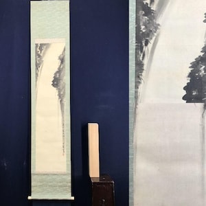 Op de afbeelding: Een verticale Japanse rolschildering met een waterval. Het kunstwerk is uitgevoerd in grijze en zwarte inkt op crèmekleurig papier, met een lichtgroene rand. De rol wordt gepresenteerd tegen een donkerblauwe achtergrond. Berglandschap.