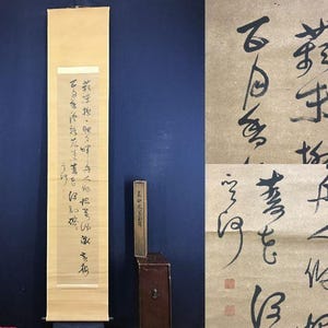 May include: An antique Asian scroll featuring black ink calligraphy on a cream-coloured background. The scroll is displayed vertically, with a wooden box to the right. The calligraphy is detailed and appears to be a poem or text.