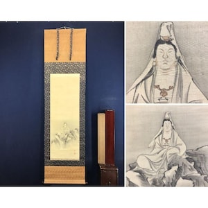 Op de afbeelding: Een traditioneel Japans rolschilderij met een zittende figuur op een berg, tegen een neutrale achtergrond. De rol heeft een bruine boven- en onderkant, met een patroonrand. Er zijn ook twee houten stukken aanwezig.