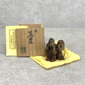 May include: Two small, brown wooden monkey figurines, one covering its ears and the other its eyes, sit on a yellow cloth. A wooden box with Japanese text and a yellow paper with Japanese text are in the background. The box is open.