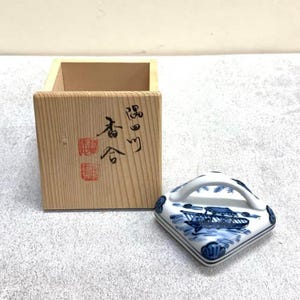 May include: A wooden box with Japanese text and a red stamp next to a square ceramic lid. The lid has a white handle and a blue and white boat design. The box and lid are on a light gray surface.