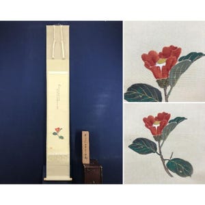 May include: A Japanese scroll painting featuring a red camellia flower with green leaves. The scroll is cream-colored with Japanese text and a decorative border. A small wooden box with text is next to the scroll.