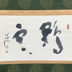 May include: A traditional Japanese calligraphy scroll featuring bold black ink characters on white paper. The scroll is framed with a wooden border and a green patterned fabric.