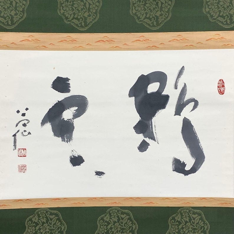 May include: A traditional Japanese calligraphy scroll featuring bold black ink characters on white paper. The scroll is framed with a wooden border and a green patterned fabric.