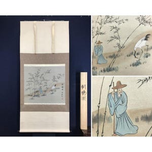 May include: A vertical hanging scroll featuring a painting of a person in a blue robe and a wide-brimmed hat standing near bamboo and a white crane. The scroll has light beige borders and includes a wooden storage box with Japanese calligraphy.