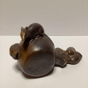 May include: A dark brown, carved wooden sculpture of a small animal, possibly a rat or mouse, perched atop a rounded form. The sculpture has a smooth, polished finish and detailed features, showcasing the artist's skill.