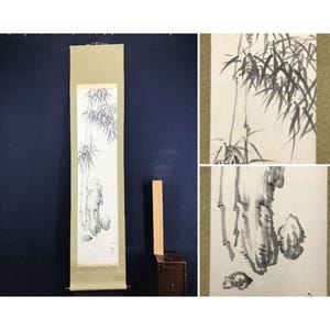 May include: A vertical hanging scroll featuring a black ink wash painting on white paper. The artwork depicts bamboo stalks, leaves, and rock formations. The scroll has a light beige fabric border with a textured pattern and a wooden rod at the top.