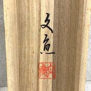 May include: Close-up of a wooden surface with visible grain. Black calligraphy and a red square stamp are on the wood. The calligraphy appears to be Asian characters. The wood has a natural, light brown color.