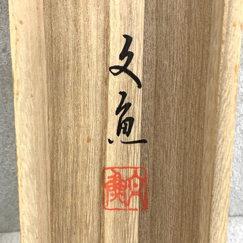 May include: Close-up of a wooden surface with visible grain. Black calligraphy and a red square stamp are on the wood. The calligraphy appears to be Asian characters. The wood has a natural, light brown color.