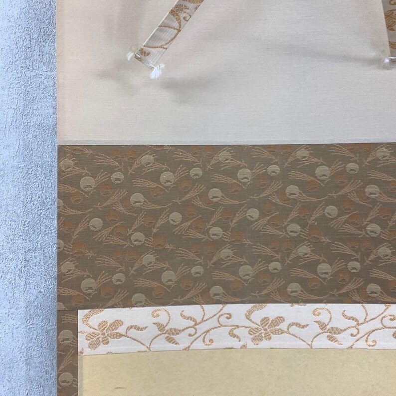 May include: Close-up of a section of a decorative scroll. The scroll features a patterned design in shades of brown and beige. A white border with a floral pattern is visible at the bottom. The scroll is held in place with clear plastic clips.