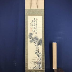 May include: A traditional Asian scroll painting featuring a detailed ink drawing of a pine tree and calligraphy. The artwork is mounted on a beige and green fabric backing, with wooden dowels at the top and bottom.