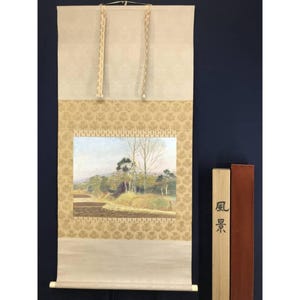 Op de afbeelding: Een Japanse rolschildering met een landschap met bomen en een pad. De rol heeft een beige achtergrond met een gouden bloemmotief en een landschapsschilderij in het midden. De tekst "landschap" staat op een houten blok.