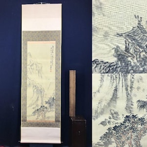 May include: An Asian scroll painting depicting a landscape with mountains, trees, and a waterfall. The artwork uses shades of grey and beige, with a detailed nature scene. The scroll is mounted on a cream-coloured backing. The painting is displayed vertically.