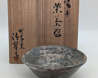 May include: A small, handcrafted ceramic bowl with a textured, mottled gray and brown surface. The bowl is set in front of a wooden box with Japanese calligraphy. The bowl has a rounded shape with a slightly flared rim.