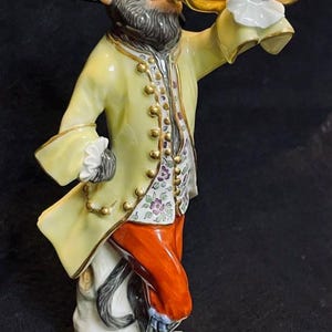 Peut inclure: Figurine en porcelaine représentant un singe vêtu d'habits du XVIIIe siècle. Le singe porte un manteau jaune avec des bordures dorées, un pantalon orange et un chapeau noir. Il tient un anneau doré, avec un col blanc à volants.