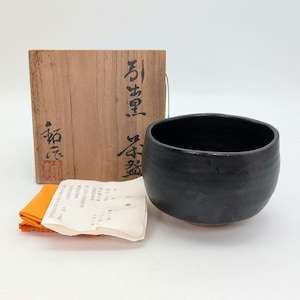 May include: A black ceramic tea bowl with a rounded shape, accompanied by a wooden storage box featuring black Japanese calligraphy and a red seal, a folded white paper document with Japanese text, and a small orange fabric cloth.