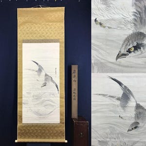 May include: A Japanese scroll painting featuring a bird in flight, rendered in shades of gray and white. The artwork is mounted on a gold-toned fabric with a decorative pattern. A wooden box with Japanese text is positioned to the side.