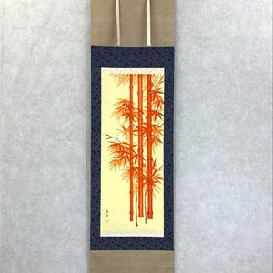 Op de afbeelding: Een verticaal Japans rolschilderij met een bamboebos in oranje en rode tinten tegen een lichtgele achtergrond. De rol is omlijst met een donkerblauwe patroonrand en een bruin top- en onderstuk.