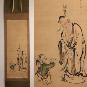 Op de afbeelding: Een antiek Japans rolschilderij met een oude man en twee kinderen. Het kunstwerk is voornamelijk in beige, groen en zwart, met gedetailleerde penseelstreken. De rol is gemonteerd op een donkergroene achtergrond.