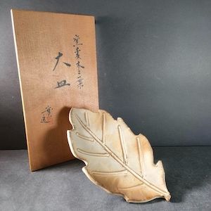 以下が含まれることがあります： 葉の形をしたセラミック皿。薄茶色の釉薬が塗られており、長方形の段ボール箱と並んでいます。箱には日本語のテキストと赤いスタンプがあります。皿には詳細な葉脈模様と質感のある表面があります。箱は薄茶色です。