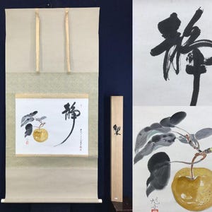 May include: Japanese scroll painting depicting a golden fruit with leaves and a calligraphic symbol. The scroll is beige with gold-coloured ties. A wooden box with Japanese characters is also present.