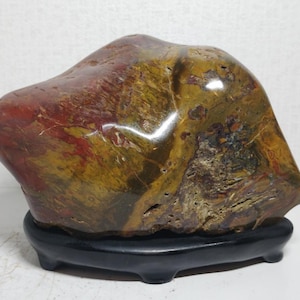 May include: A polished, natural stone sculpture with a smooth, rounded form. The stone features a blend of colors, including reddish-brown, yellow, and hints of green. It rests on a small, black, four-legged stand, showcasing the stone's unique patterns.