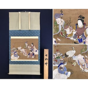 May include: Japanese scroll painting featuring figures, cranes, and other elements. The artwork is mounted on a light green and blue fabric. A wooden sign with Japanese characters is placed next to the scroll.