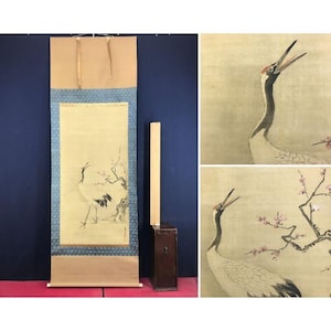May include: A Japanese scroll painting featuring a crane with its beak open, standing near a flowering branch. The artwork is framed with a blue patterned border and a tan top. A wooden box and a long wooden piece are next to the scroll.