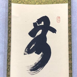 May include: A traditional Japanese calligraphy scroll featuring a bold, abstract black ink character. The artwork is set against a cream-colored background, framed by a decorative green and gold border. A red seal is visible in the upper right corner.