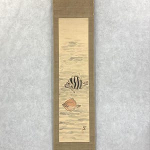 May include: A long, vertical scroll featuring a watercolor painting of two fish. One fish is black and white striped, the other is orange. The background depicts water with light brown and gray tones. The scroll is framed with a brown border and wooden dowels.