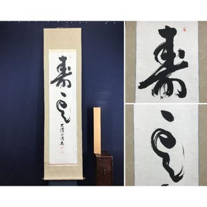 May include: A Japanese calligraphy scroll with striking black brushstrokes on white paper. The artwork features several characters and symbols, with a wooden dowel at the bottom. The scroll is set against a dark blue backdrop.